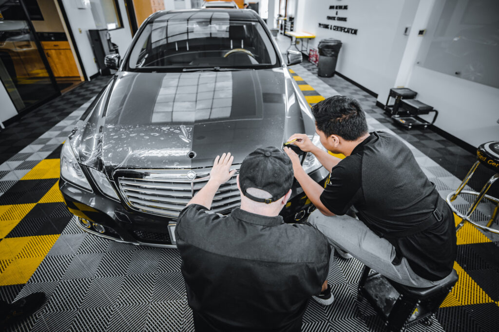 Two men receive paint protection film training while working on a car in a garage.