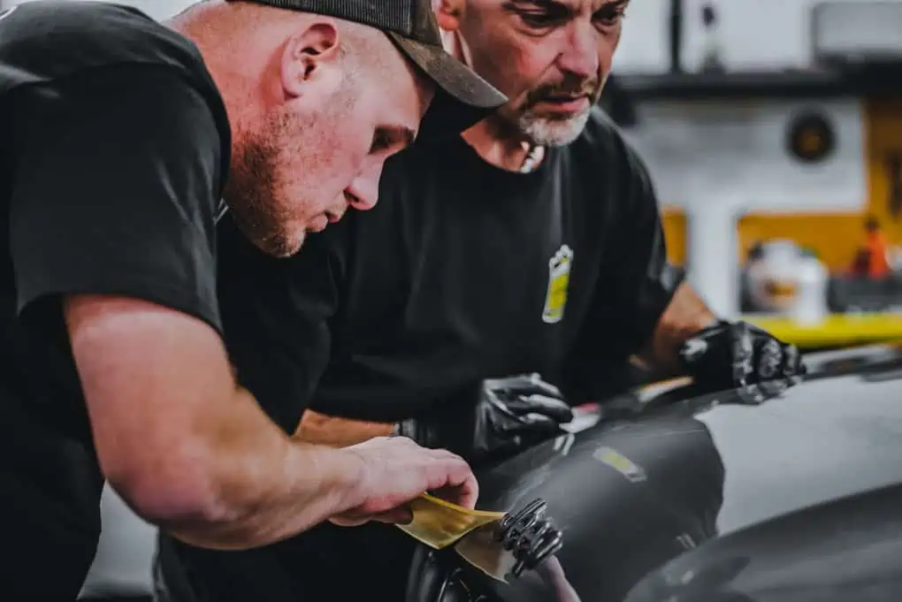Two men receiving paint chip repair training while working on the hood of a car.