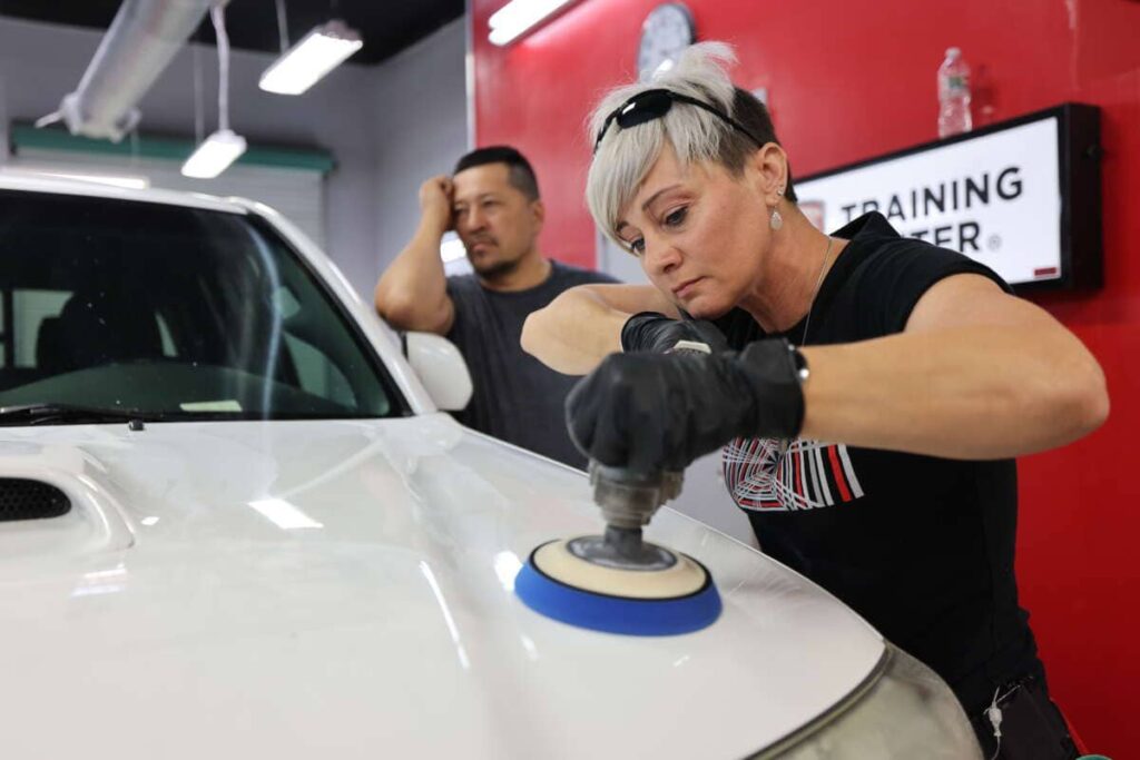 A woman is receiving paint correction training by polishing a white car in a shop.