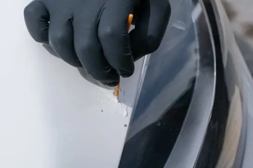 A person with black gloves is using a tool to perform paint chip repair on a white car.