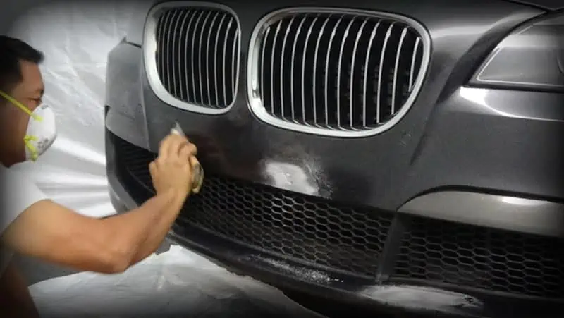 A man is learning scuffed bumper repair training techniques by spraying a black BMW while wearing a mask.