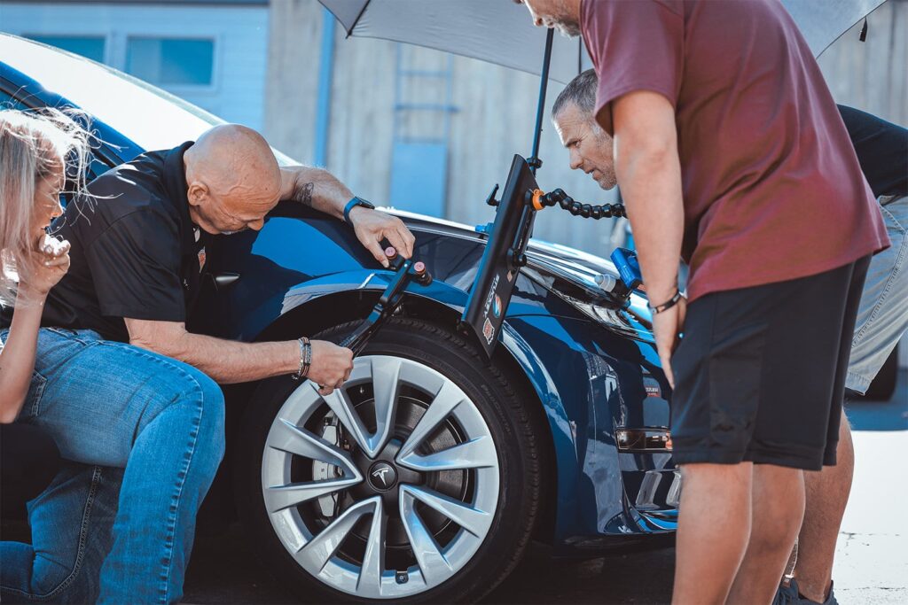 Two men inspect the front left tire of a blue car, with one woman observing from the side. An umbrella provides shade as they discuss Ceramic Coating Training techniques.