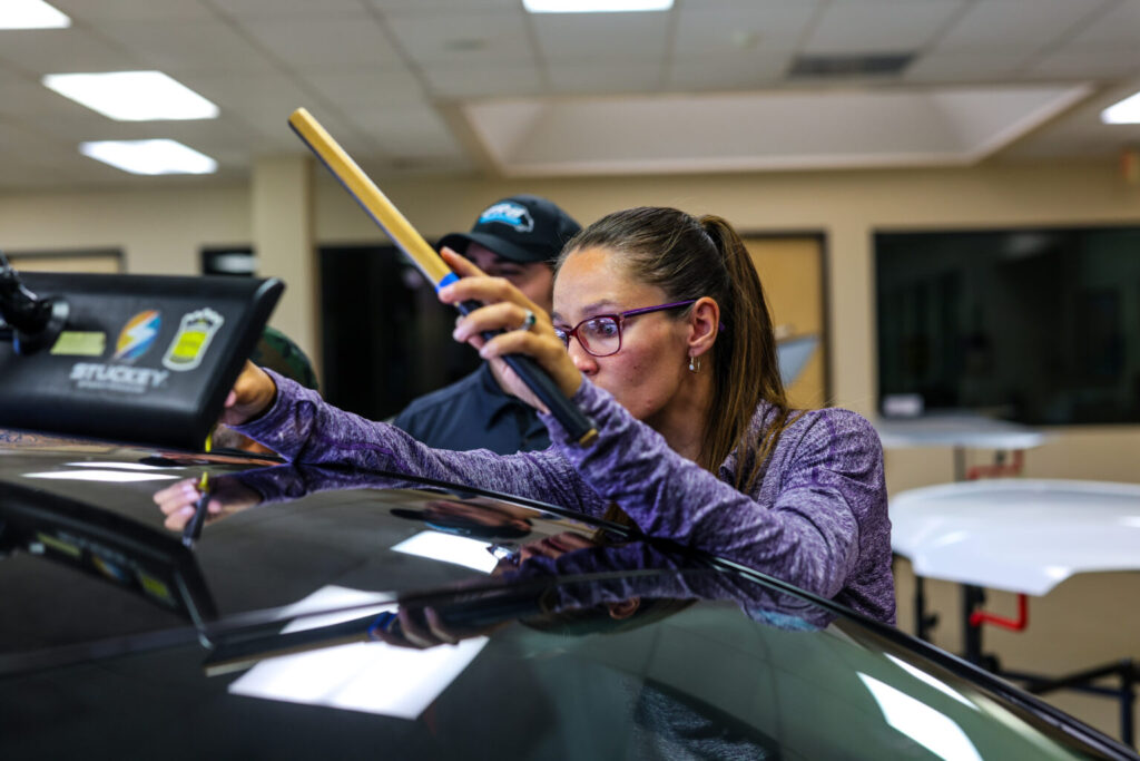 A person wearing glasses and a purple jacket uses a wooden tool to work on the roof of a black car in an indoor setting during a ceramic coating training session.