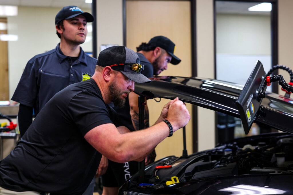 Three mechanics in a workshop working on a car with its hood open. One focuses on the engine using tools, while the others observe and assist. They are discussing the benefits of ceramic coating training to enhance their skills and improve overall vehicle performance.