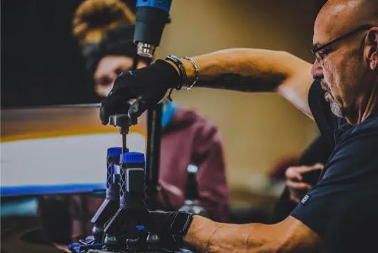 A person wearing gloves operates a machine tool, likely undergoing Ceramic Coating Training, accompanied by another individual in the background.