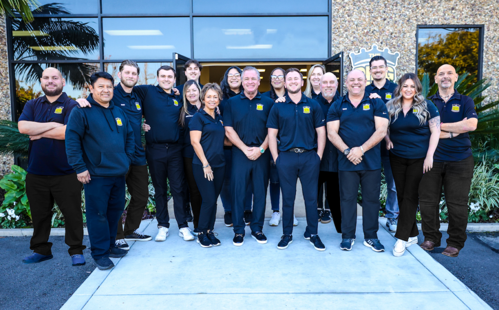 A group of 15 people stand in front of a building entrance, all wearing navy blue uniforms with logos. The group, participating in Ceramic Coating Training, consists of both men and women posing together for the photo.