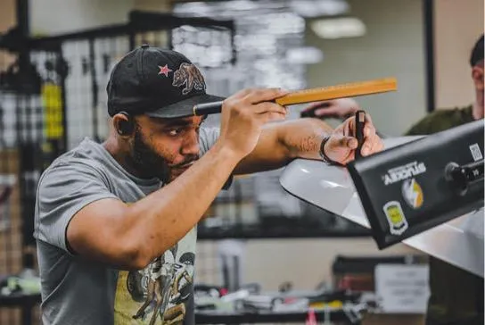 A man wearing a cap and a t-shirt is meticulously measuring something with a ruler next to a computer monitor, likely preparing for his Ceramic Coating Training.