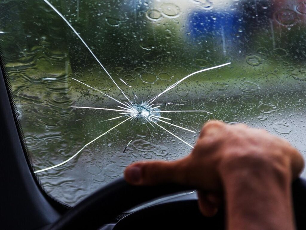 A cracked windshield with a spiderweb pattern is pictured from inside a car. The driver's hand is on the steering wheel, rain visible on the glass. This scenario highlights the importance of Paint Protection Film Training to safeguard vehicles against such damage.