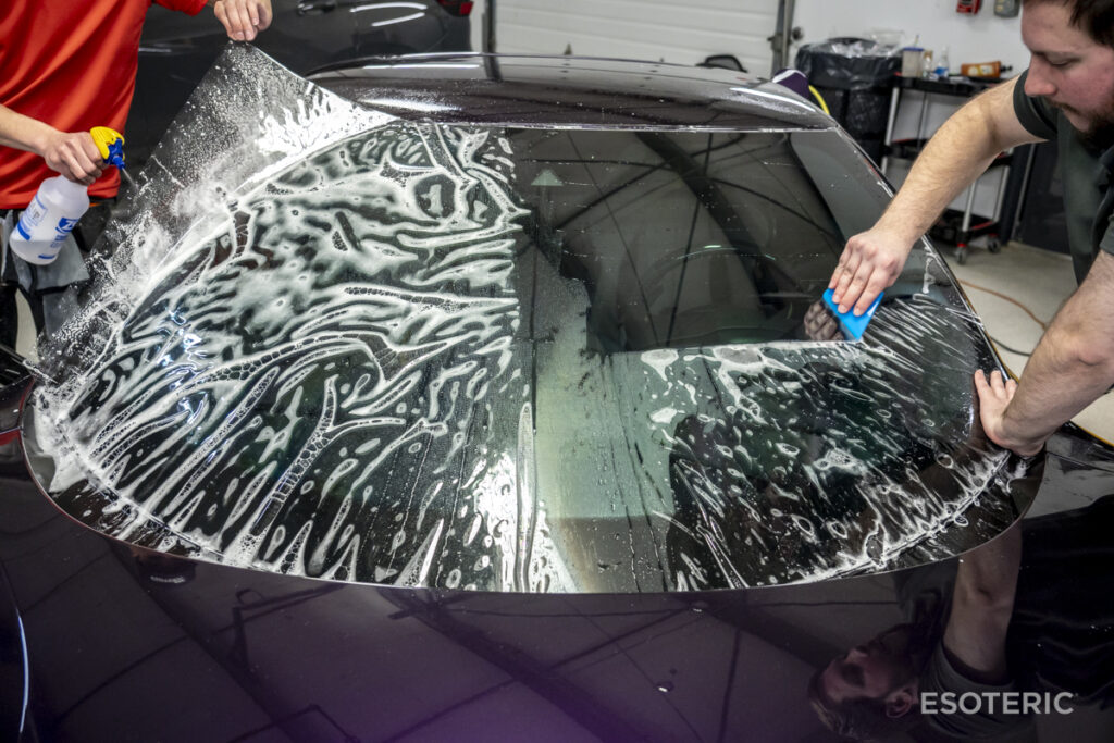 Two individuals undergo paint protection film training as they apply a tinted protective film to the rear windshield of a black car in a garage, using smoothing tools and a spray bottle.