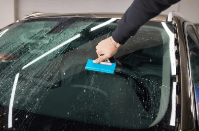 A person is using a blue squeegee to clean the exterior of a car's windshield, which is covered in soapy water, ensuring it's pristine for an upcoming Paint Protection Film Training session.