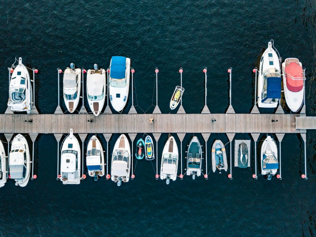 An aerial view of multiple boats docked alongside a wooden pier, with a person walking on the pier. The boats vary in size and type, arranged in an orderly fashion, reminiscent of the precision seen in Paint Protection Film Training.