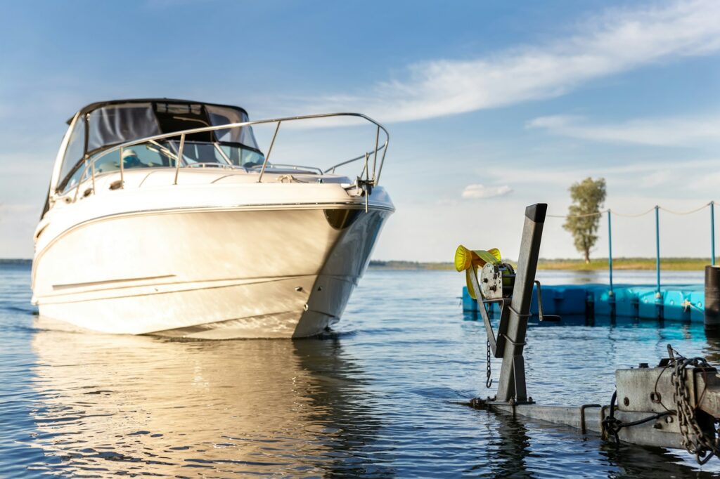 A white motorboat, sporting the latest paint protection film training decals, is partially out of the water next to a boat dock on a sunny day, with a visible anchor and calm waters.