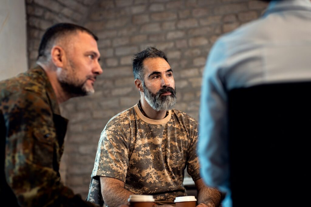 Three men having a conversation while sitting in a room with exposed brick walls; two of them are wearing camouflage attire and discussing ceramic coating training over coffee cups on the table.