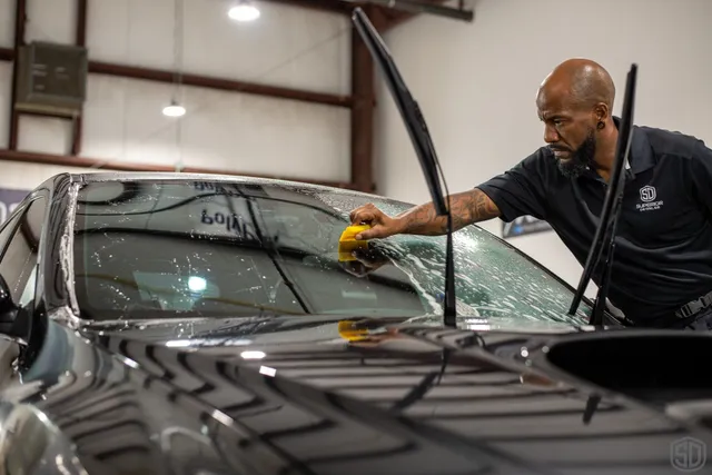 A person is cleaning the windshield of a black car with a yellow sponge in a well-lit indoor setting, meticulously preparing the surface as if it were part of Paint Protection Film Training.