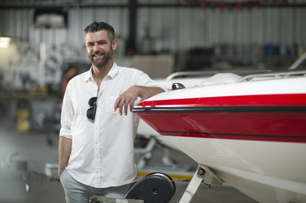 A man with a beard and wearing a white shirt stands beside a red and white boat in what appears to be an indoor facility, possibly used for Paint Protection Film Training. He has one hand in his pocket and is smiling at the camera.