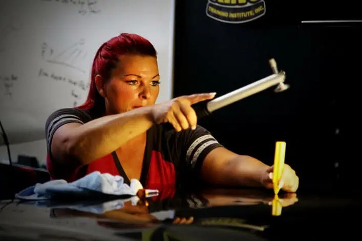 A woman with red hair uses a tool to fix a car's surface. She wears a red and black shirt, and a towel is placed on the car's hood. A whiteboard in the background has diagrams and notes.