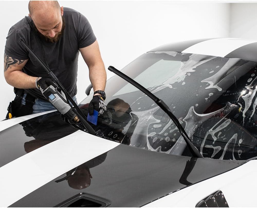 A person meticulously applies a tinted film on a car windshield, using expert squeegee techniques and a heat gun. Thanks to comprehensive paint protection film training, every move is precise, ensuring flawless installation and maximum durability.