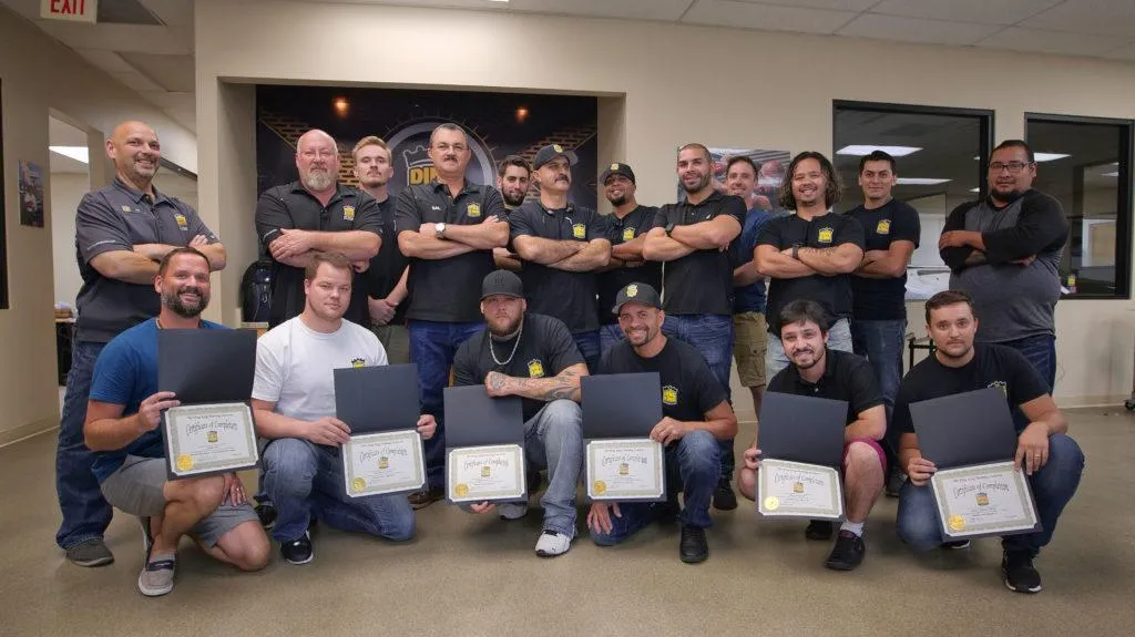 A group of 16 men, some standing and some kneeling, pose indoors after completing their Paint Protection Film Training. Eight in front proudly hold certificates. They're in a room with beige walls and windows.