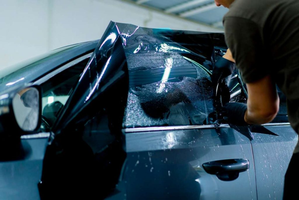 Inside the garage, a person deftly applies tinted film to a car window, showing skill reminiscent of paint protection film training.