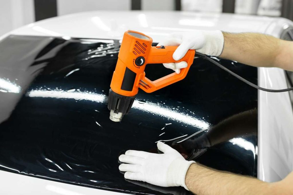 A person, having completed paint protection film training, uses a heat gun to apply tinted film to a car window, carefully wearing white gloves for safety.