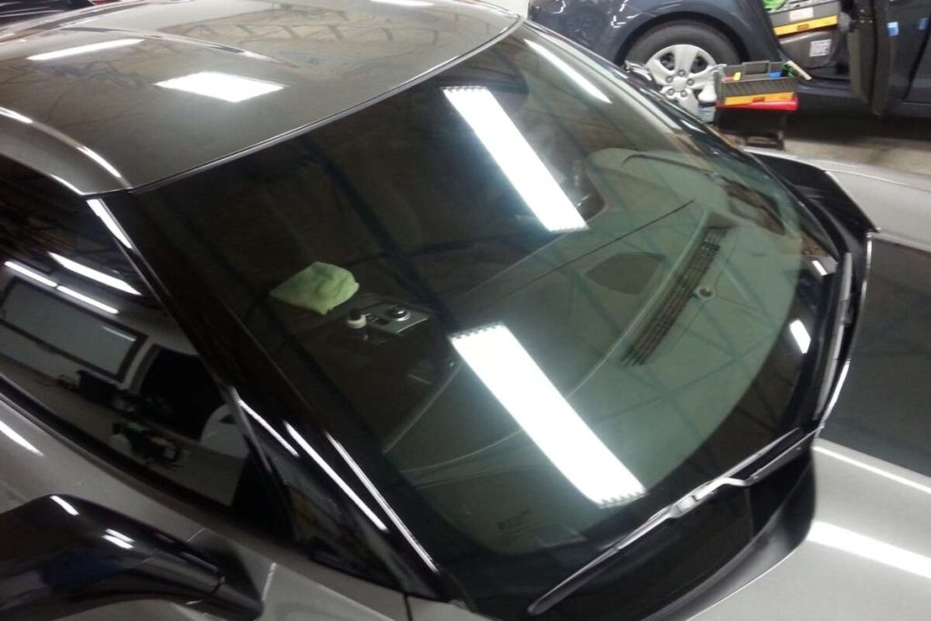 Close-up of a car windshield reflecting overhead lights in a garage. A cloth rests on the dashboard, hinting at recent Paint Protection Film training. Another car is visible in the background, showcasing pristine attention to detail.