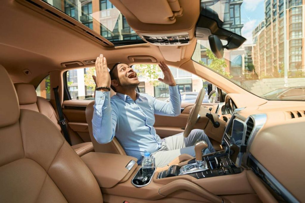 A man in a blue shirt, fresh from paint protection film training, sits in a car with a beige interior, looking upward with an expression of frustration and his hands raised.