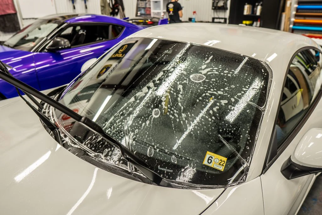 A gray car with its wiper raised is getting a thorough windshield cleaning with soap and water, while a purple car, possibly awaiting paint protection film training, is parked nearby inside the garage.