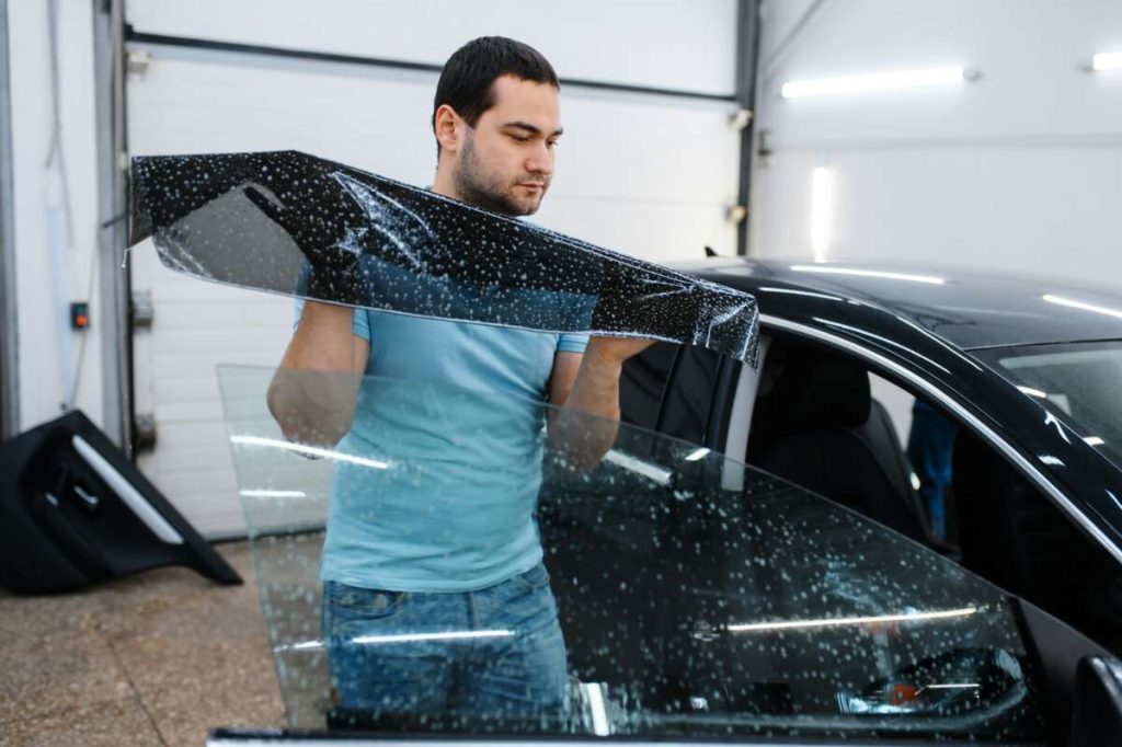 In a garage bathed in soft light, a man skillfully applies tinted film to a car window, harnessing techniques from his paint protection film training.