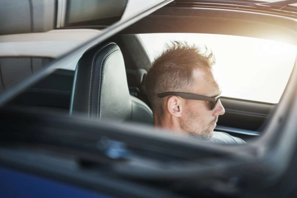 A man wearing sunglasses navigates the road, his car gleaming from a recent paint protection film training session, captured through the window at a side angle with sunlight casting a warm glow in the background.