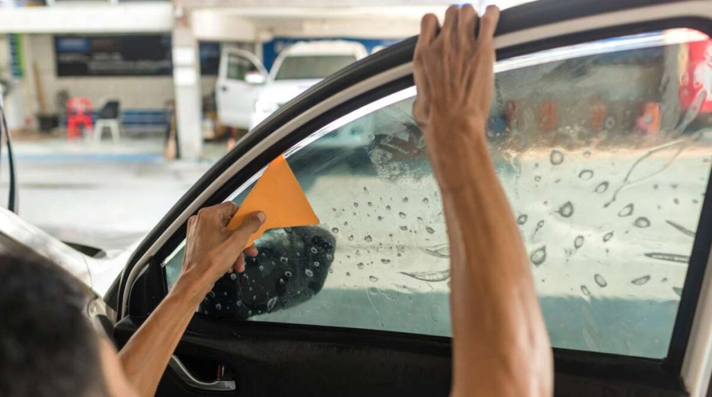 Applying tint film to a car window using an orange tool, with water droplets glistening on the glass surface, requires precision. Mastering this skill is a crucial part of Paint Protection Film Training.