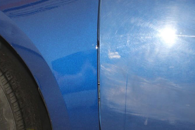 Close-up of a blue car showing a slight misalignment between the front panel and door, with a reflection of light visible on the surface.